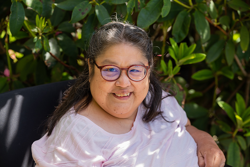 A photo of Charlene who is wearing glasses and smiling while sitting in front of a large outdoor plant.