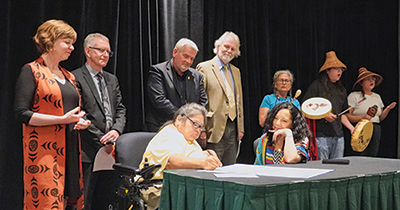 A group of government and CLBC leaders stand next to a table watching while Charlene signs a reconciliation commitment document. In the background is a group of three Indigenous drummers .