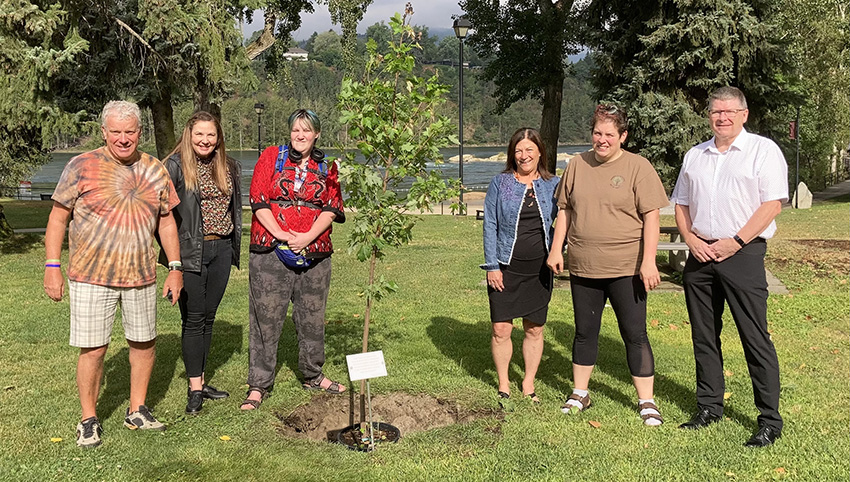 A group of 6 men and women stand together next to a newly planted tree. There is a plaque in front of the tree.