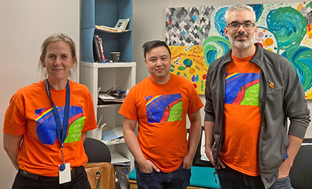 Three CLBC staff members stand together and wear orange shirts with an artwork named "Every Child Matters" on the front of the shirts.