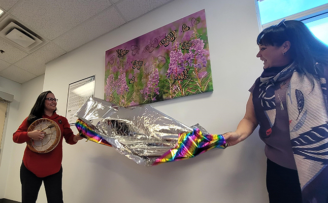 Two women remove a cover, unveiling an Indigenous artwork hanging on a wall.