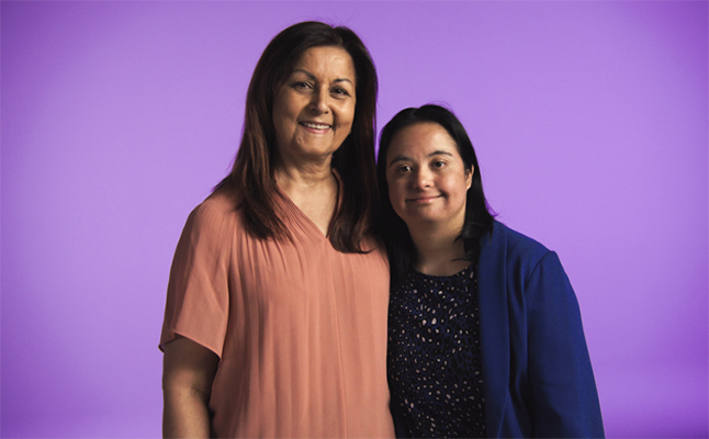 Two women, a mother and daughter, stand side by side with an arm around each other smiling in front of a purple background.